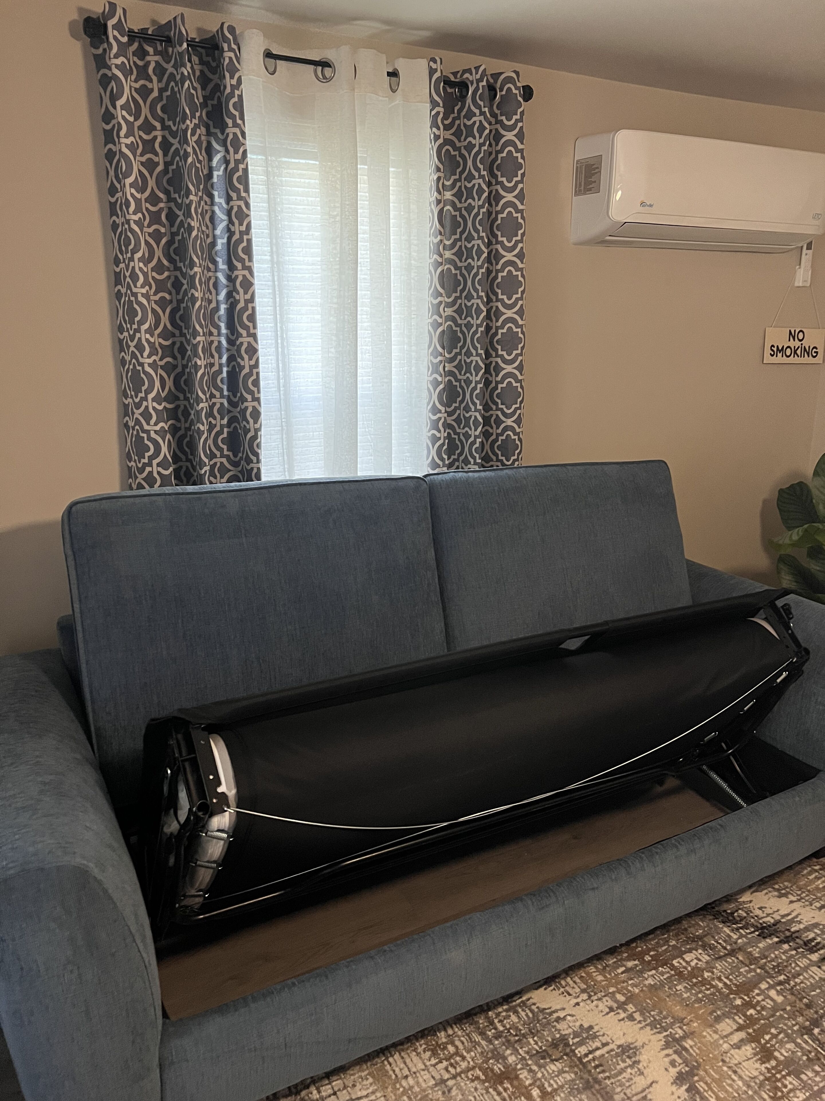 A living room with a grey sofa, a black storage unit, patterned curtains, a window with sheer white curtains, an air conditioning unit, and a 'No Smoking' sign.