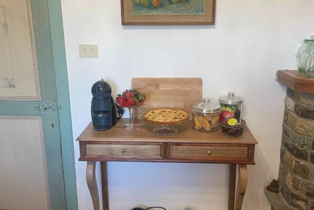 A small kitchen with a wooden table, a coffee maker, a pie, a jar of jam, and a fruit basket.