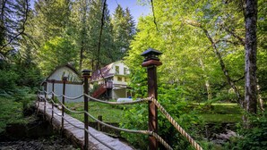 Eine hölzerne Brücke mit Seilgeländern führt zu einem Haus, das von dichtem Wald umgeben ist.
