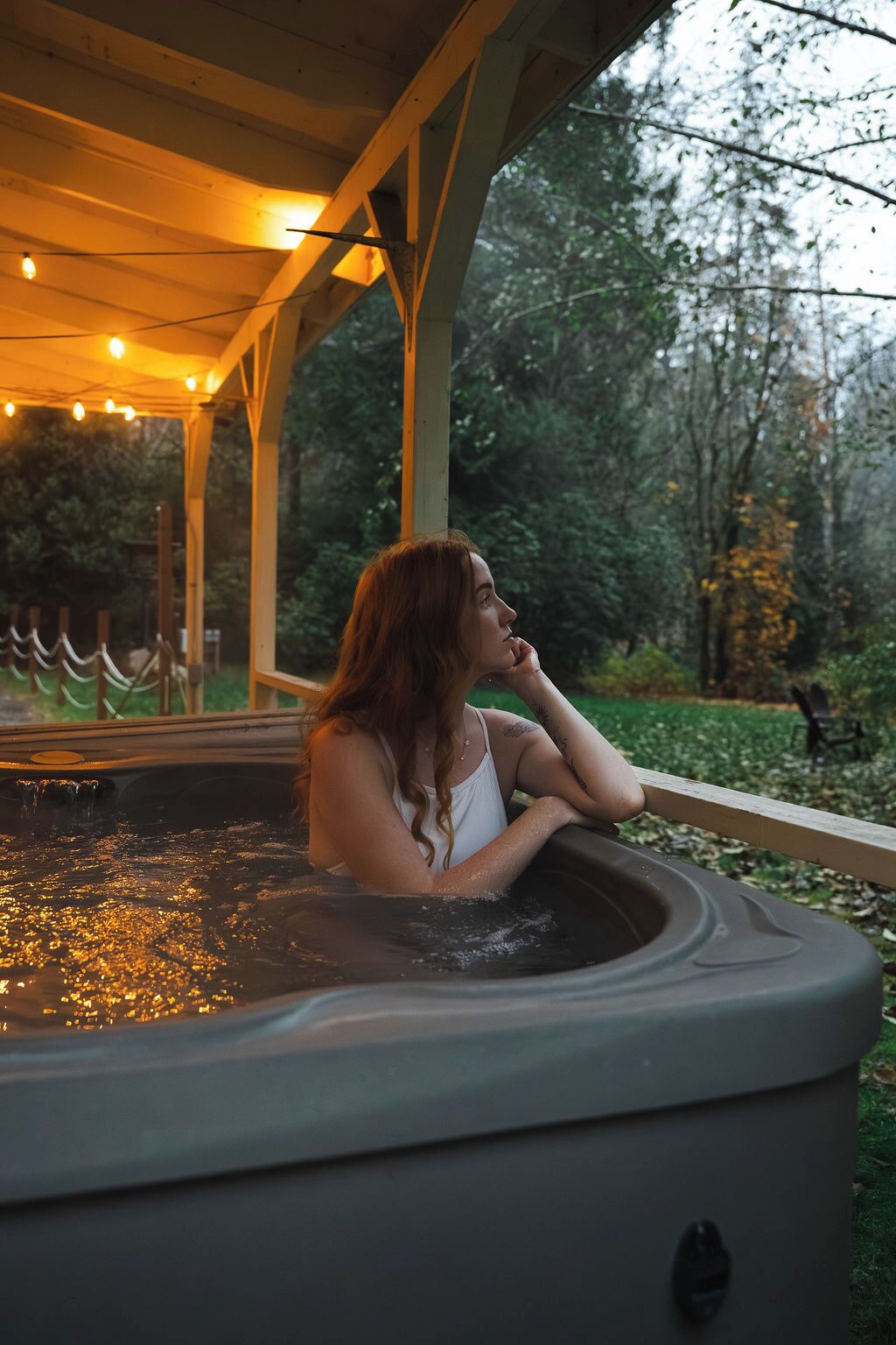 A person relaxing in a hot tub outdoors, with a wooden deck and greenery in the background.