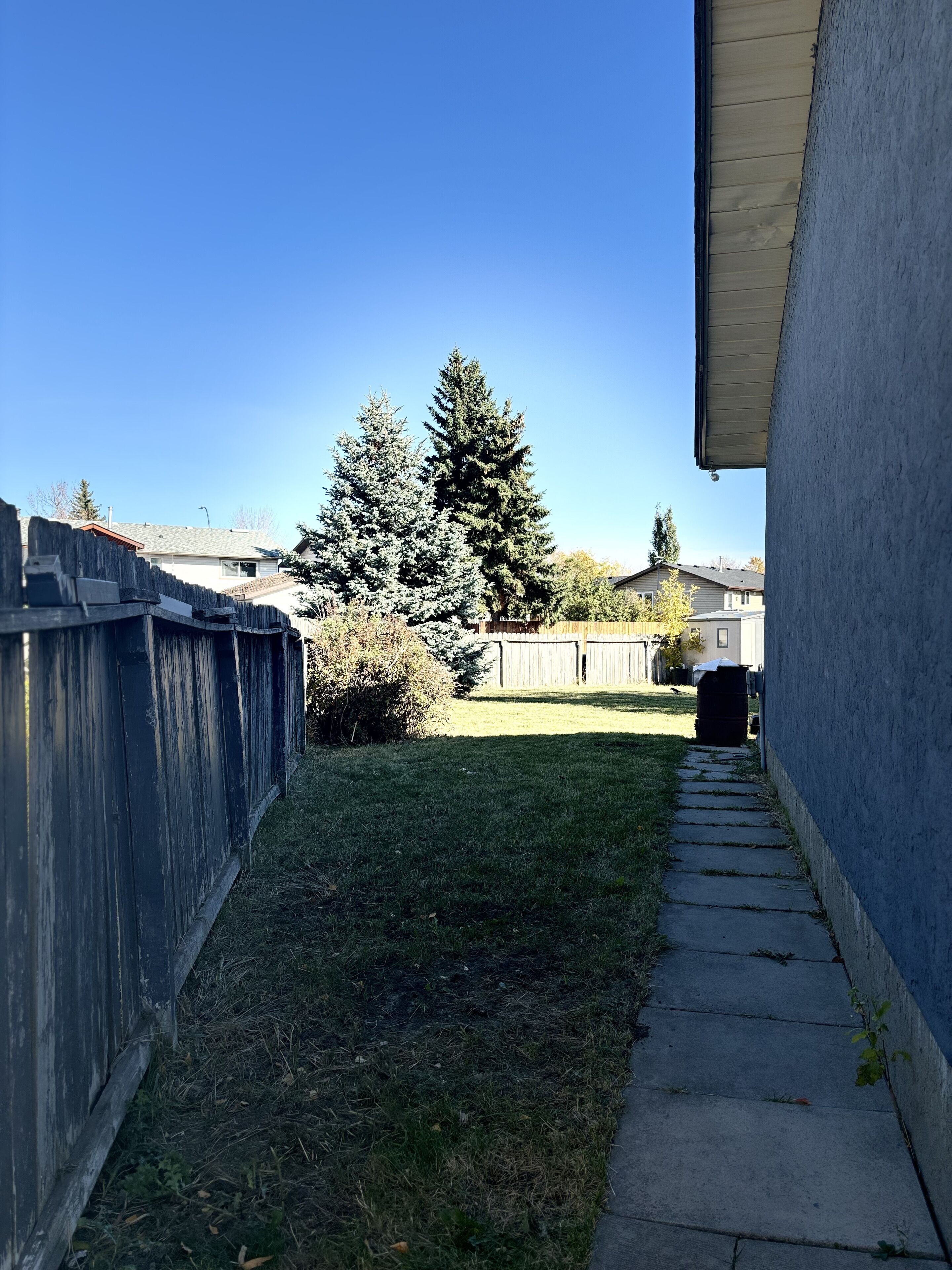 Un étroit sentier en béton mène à une zone herbeuse, bordée d’une clôture en bois d’un côté et d’un bâtiment de l’autre.