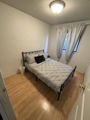 A bedroom with a bed, a bedside table, a window with curtains, and a ceiling light. - Welcome to Falcon Escape (Calgary)
