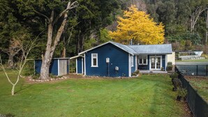 A blue house with a porch, a small shed, and a large tree with yellow leaves. - Fantail Cottage Kaiuma Bay - Havelock Holiday Home (Havelock)