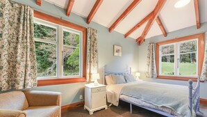 A bedroom with a bed, nightstand, chair, and large windows with curtains. - Fantail Cottage Kaiuma Bay - Havelock Holiday Home (Havelock)
