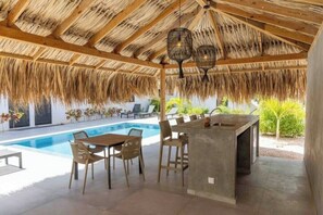 Un espace piscine extérieur doté d’un toit de chaume, d’un bar et de coins salon.