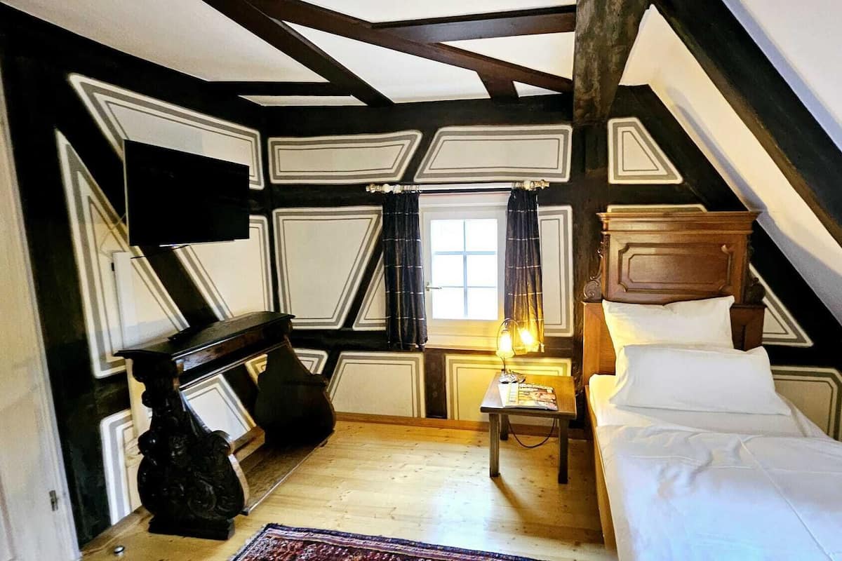 Een kamer met een houten plafond, een bed, een nachtkastje, een lamp en een raam met gordijnen.