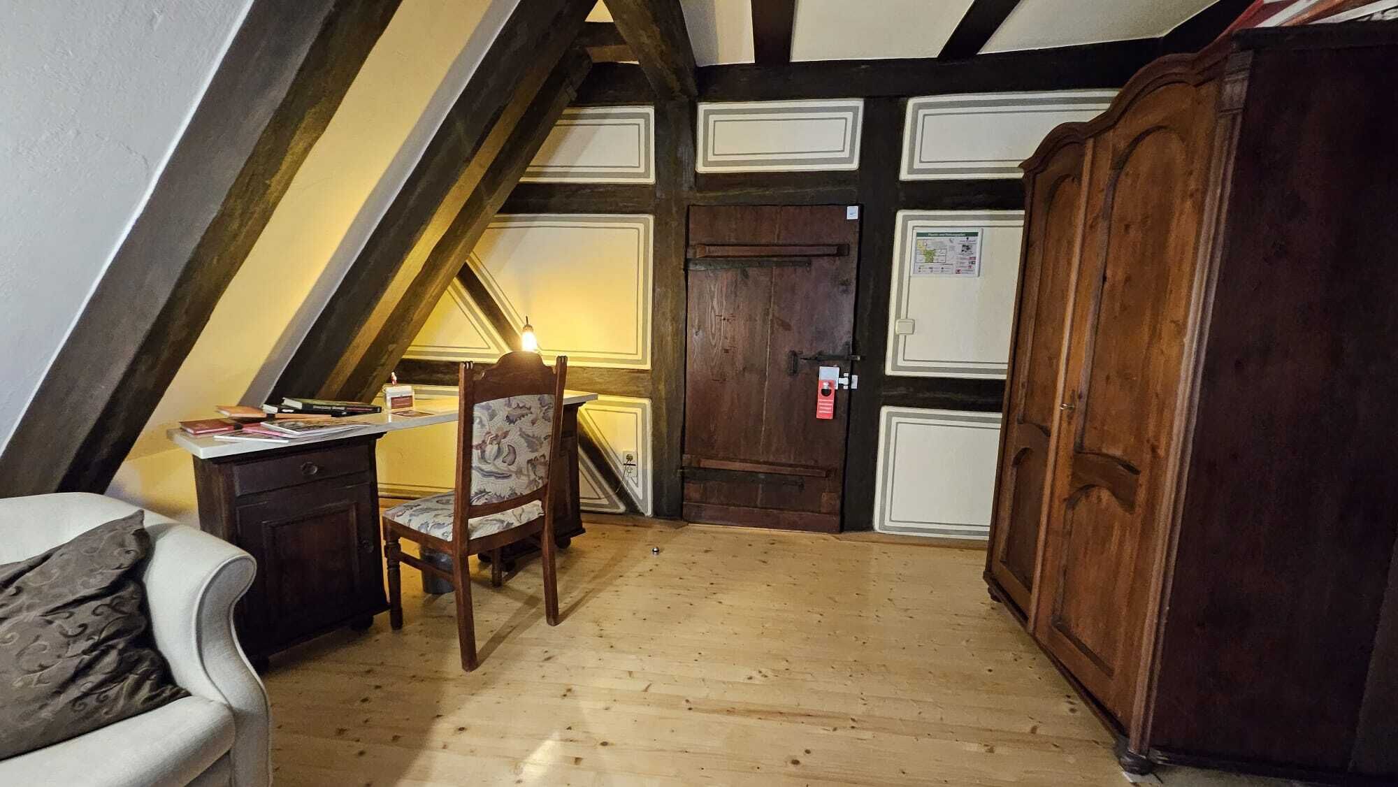 A room with a sloped ceiling, wooden floor, a desk with a chair, a wardrobe, and a door with a notice.