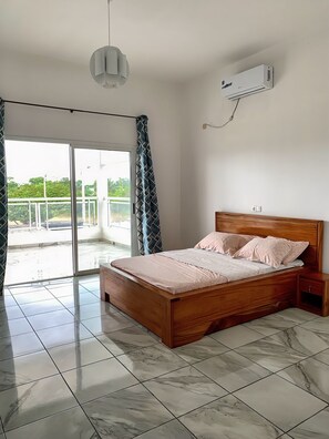 Een slaapkamer met een houten bed, nachtkastje, een wandairconditioner en uitzicht door een glazen schuifdeur op een buitenbalkon.