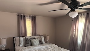 Ein Schlafzimmer mit Bett, Deckenventilator, Nachttisch mit Lampe und Fenster mit Vorhängen.