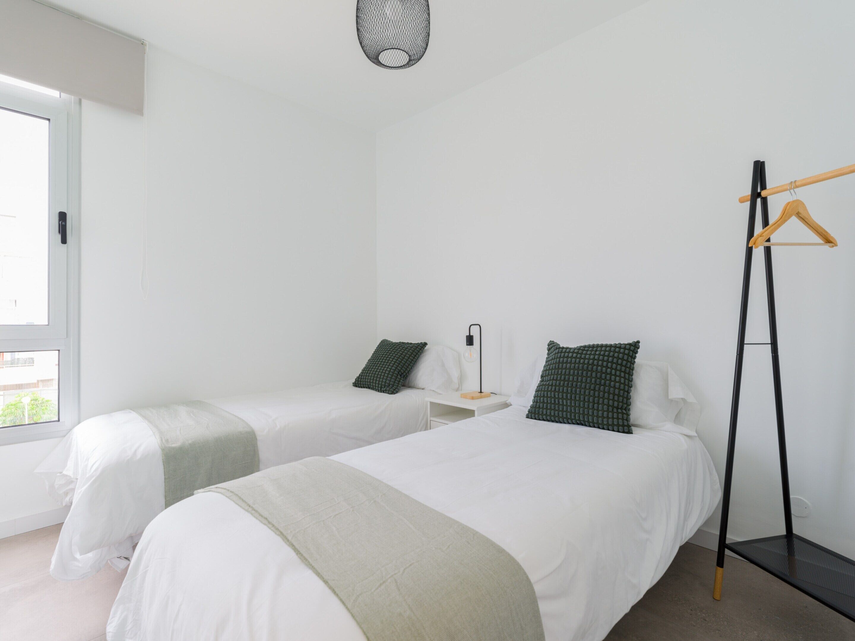 A hotel room with two single beds, a bedside table with a lamp, a hanging clothes rack, and a window with a view.