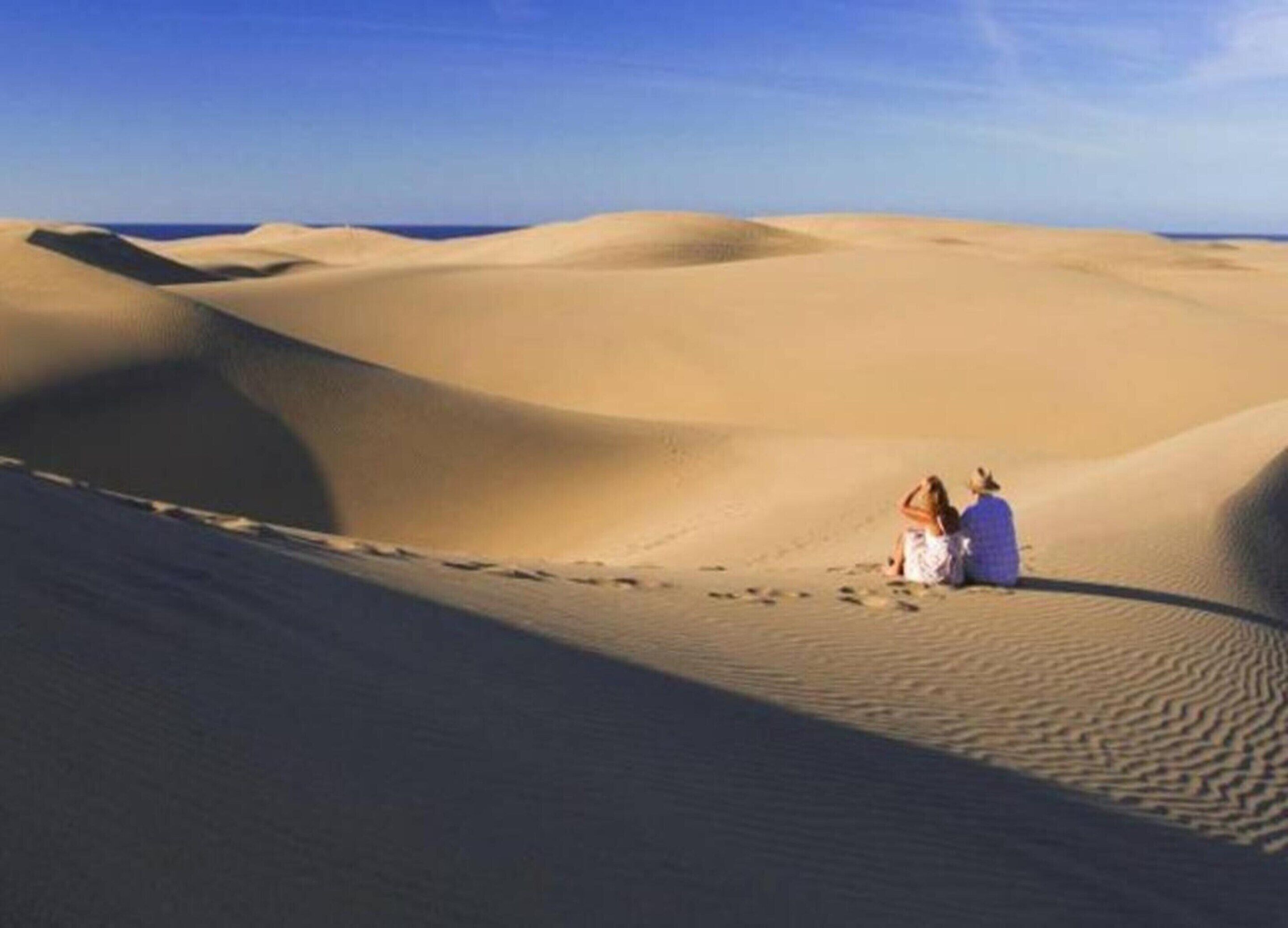 A desert landscape with sand dunes and two people sitting on the sand.