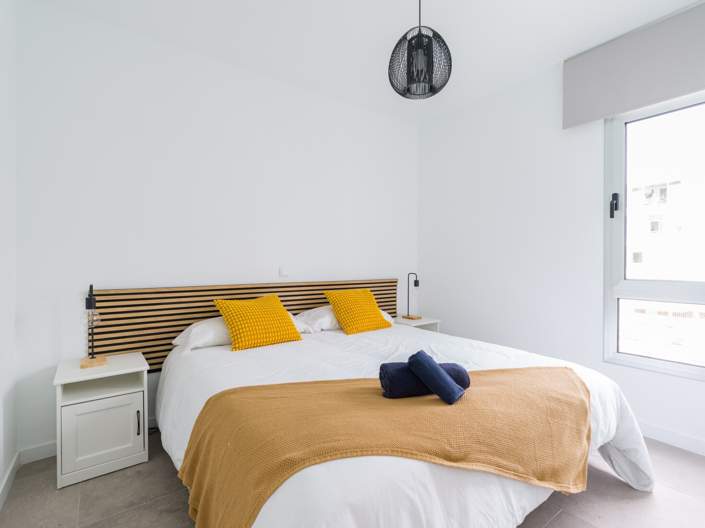 A modern bedroom with a wooden headboard, two bedside tables, a large window, and a hanging pendant light.