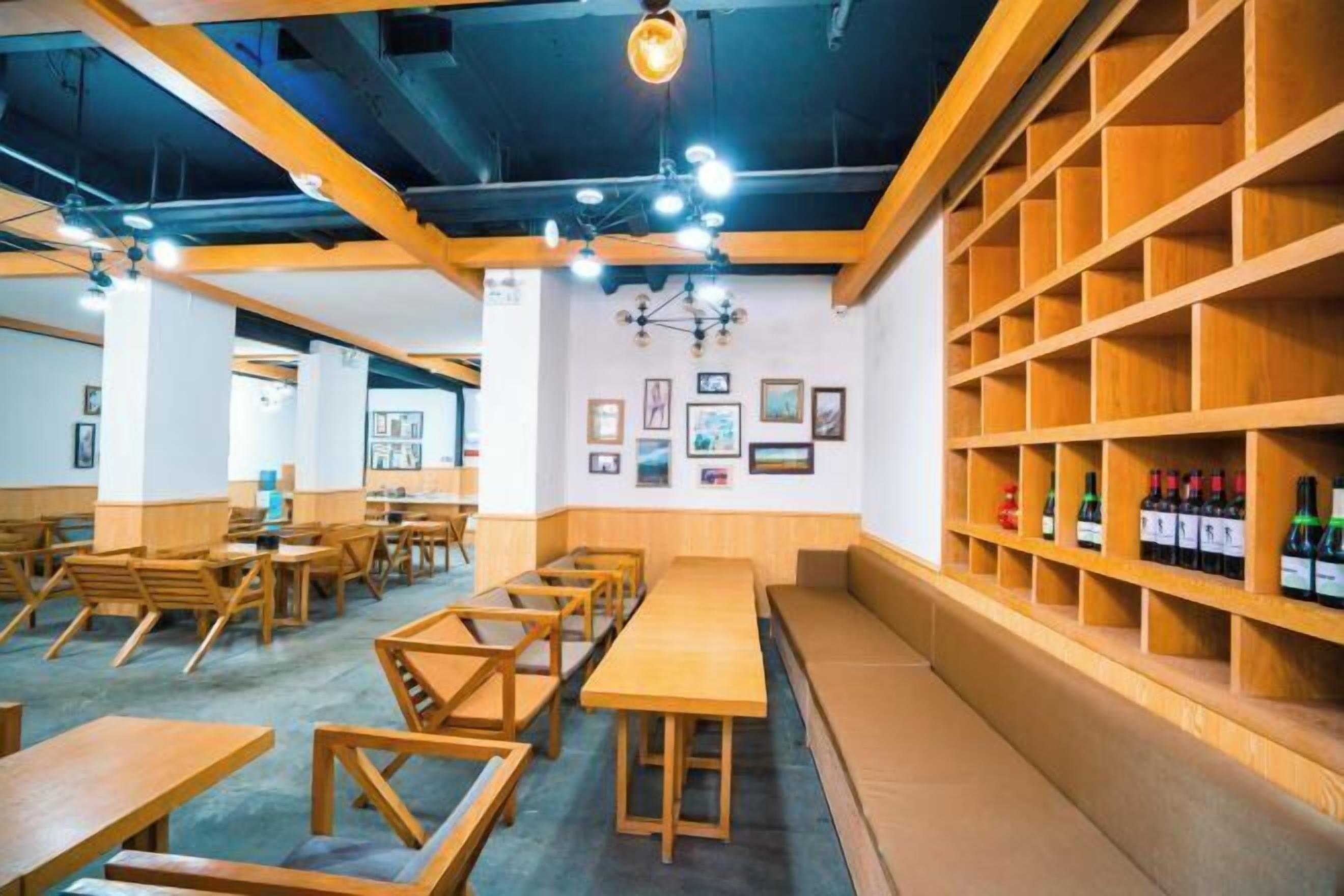 A café with wooden tables and chairs, a wall with framed pictures, and shelves with bottles.