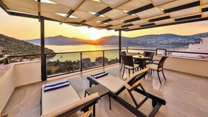 Μια βεράντα με τραπέζι και καρέκλες, από την οποία μπορεί κανείς να απολαύσει την υπέροχη θέα στη θάλασσα και τα βουνά κατά τη διάρκεια του ηλιοβασιλέματος.