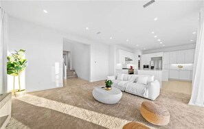 Contemporary living space with a sofa, ottoman, and potted plant, opening to a kitchen with white cabinetry and stainless steel appliances.