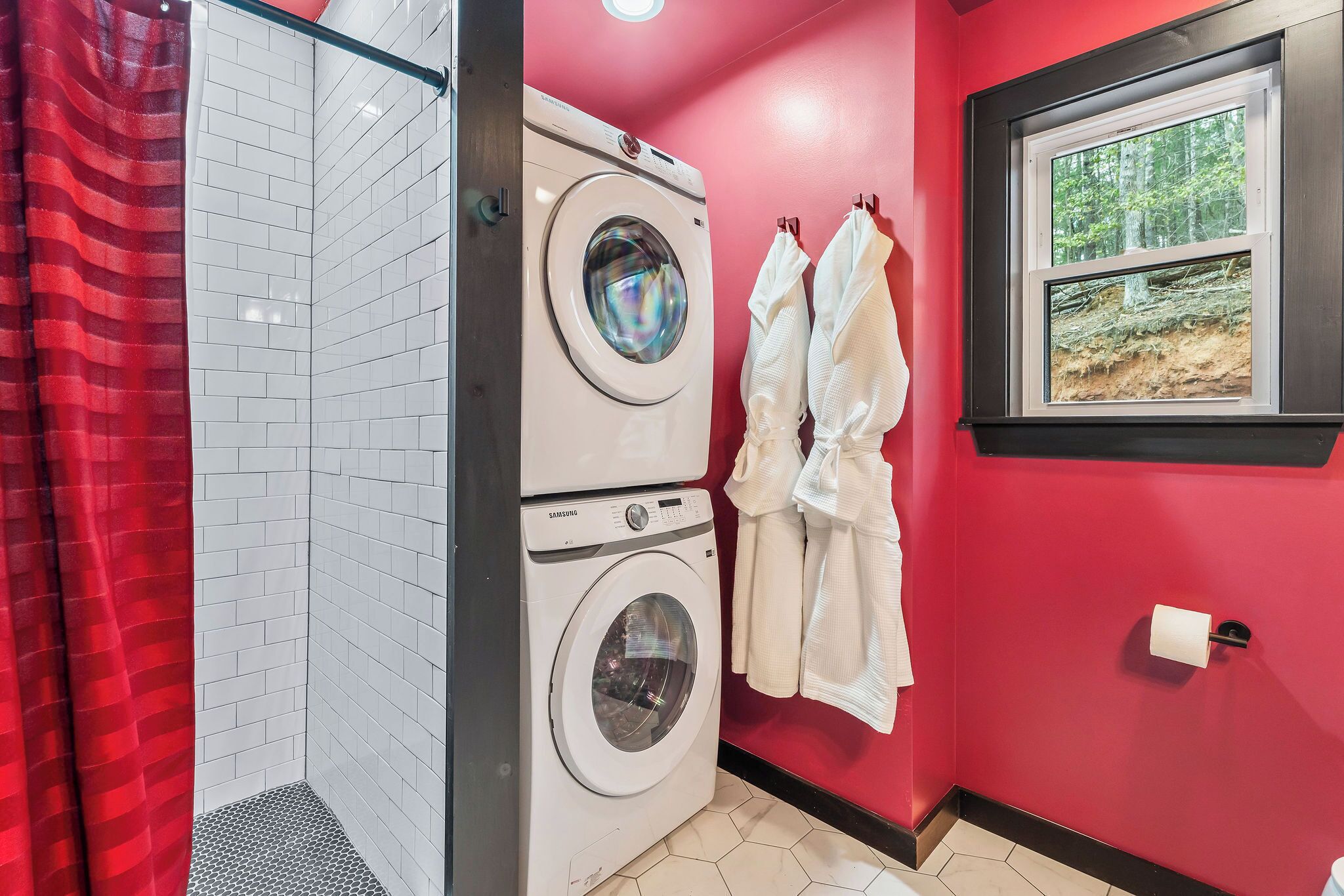 A laundry room with a stacked washer and dryer, a red shower curtain, and a window with a forest view.