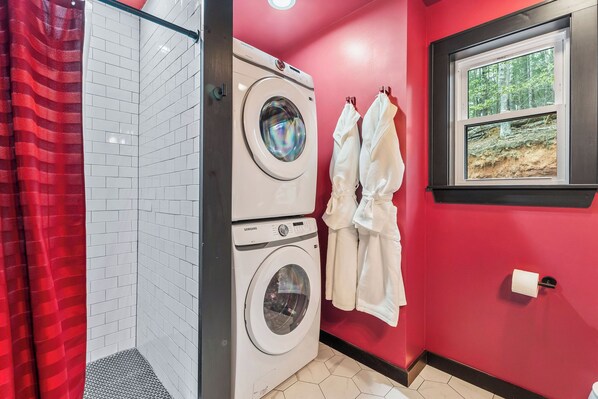 A laundry room with a stacked washer and dryer, a red shower curtain, and a window with a forest view.