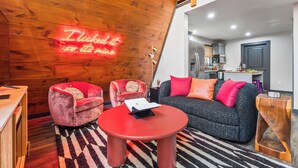 A modern living room with a red table, two red armchairs, and a black sofa with orange cushions. A neon sign on the wall reads "I tickled it so it came."