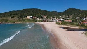A coastal beach with clear waters, a sandy shore, and a backdrop of hills with buildings. - Contêiner Exclusivo - Praia da Gamboa (Garopaba)