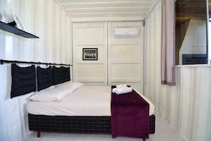 A neatly made bed with a black headboard, a white mattress, and a purple throw blanket. There is a framed picture on the wall above the bed. - Contêiner Exclusivo - Praia da Gamboa (Garopaba)