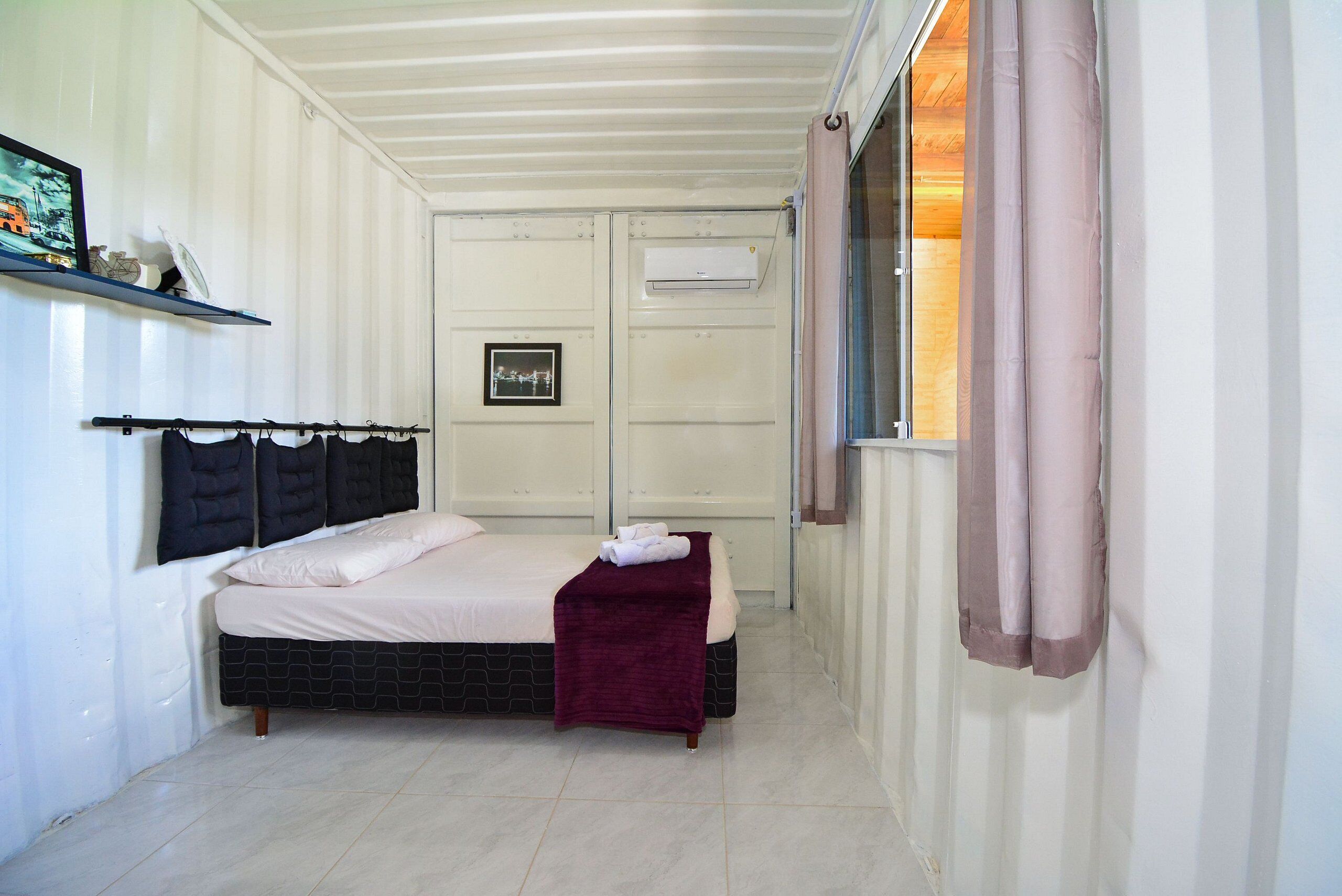 Quarto com cama, pequena mesa de cabeceira, prateleira na parede, quadro emoldurado e janela com cortinas.
