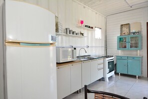 A kitchen with white cabinets, a microwave, a sink, and a stove. There are shelves with various items and a teal cabinet with glass doors. - Contêiner Exclusivo - Praia da Gamboa (Garopaba)