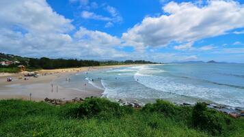 Uma praia com céu claro e ondas, uma faixa de areia e vegetação ao primeiro plano.