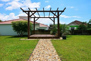 Un jardin agrémenté d’une pergola en bois, d’un chemin de gravier et de maisons en arrière-plan.