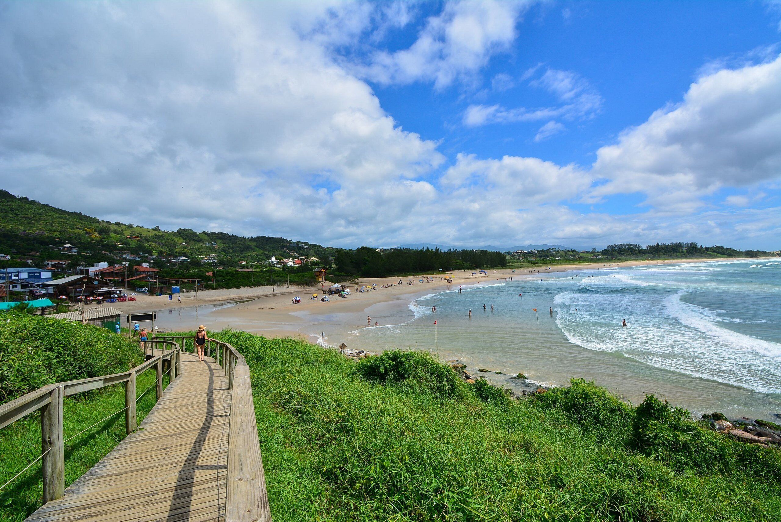 A beach with a wooden boardwalk leading to the shore, surrounded by greenery and houses in the background.