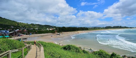 A beach with a wooden boardwalk leading to the shore, surrounded by greenery and houses in the background.