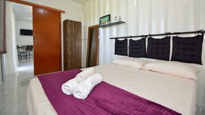 Ein Hotelzimmer mit einem Bett, einer violetten Decke, zwei aufgerollten Handtüchern, einer Holztür, einem Fernseher auf einem Regal und einem kleinen Tisch mit Stühlen.