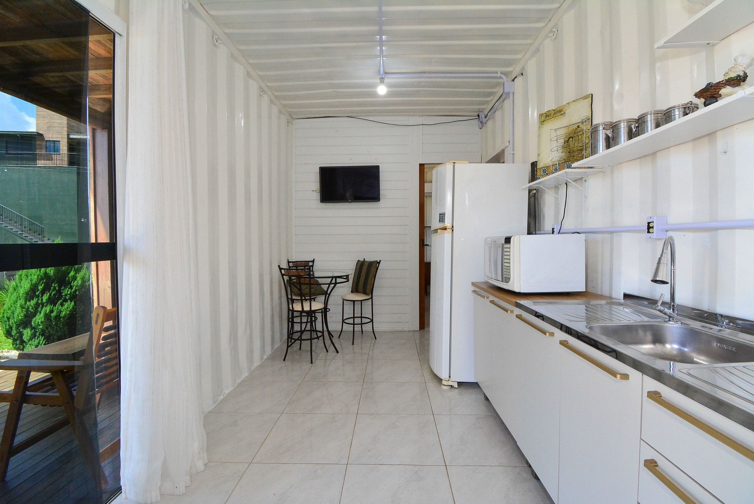 Cozinha compacta com armários brancos, geladeira, micro-ondas e pia. Possui um pequeno espaço de refeições com duas cadeiras e uma mesa.