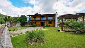A two-story building with a yellow facade, a balcony, and a well-maintained lawn with palm trees. - Contêiner Exclusivo - Praia da Gamboa (Garopaba)