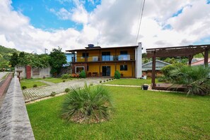 A two-story building with a yellow facade, a balcony, and a well-maintained lawn with palm trees. - Contêiner Exclusivo - Praia da Gamboa (Garopaba)