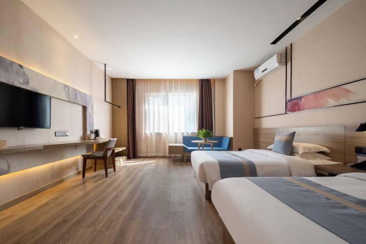 A hotel room with a large bed, a desk, a chair, a TV mounted on the wall, and a window with curtains.