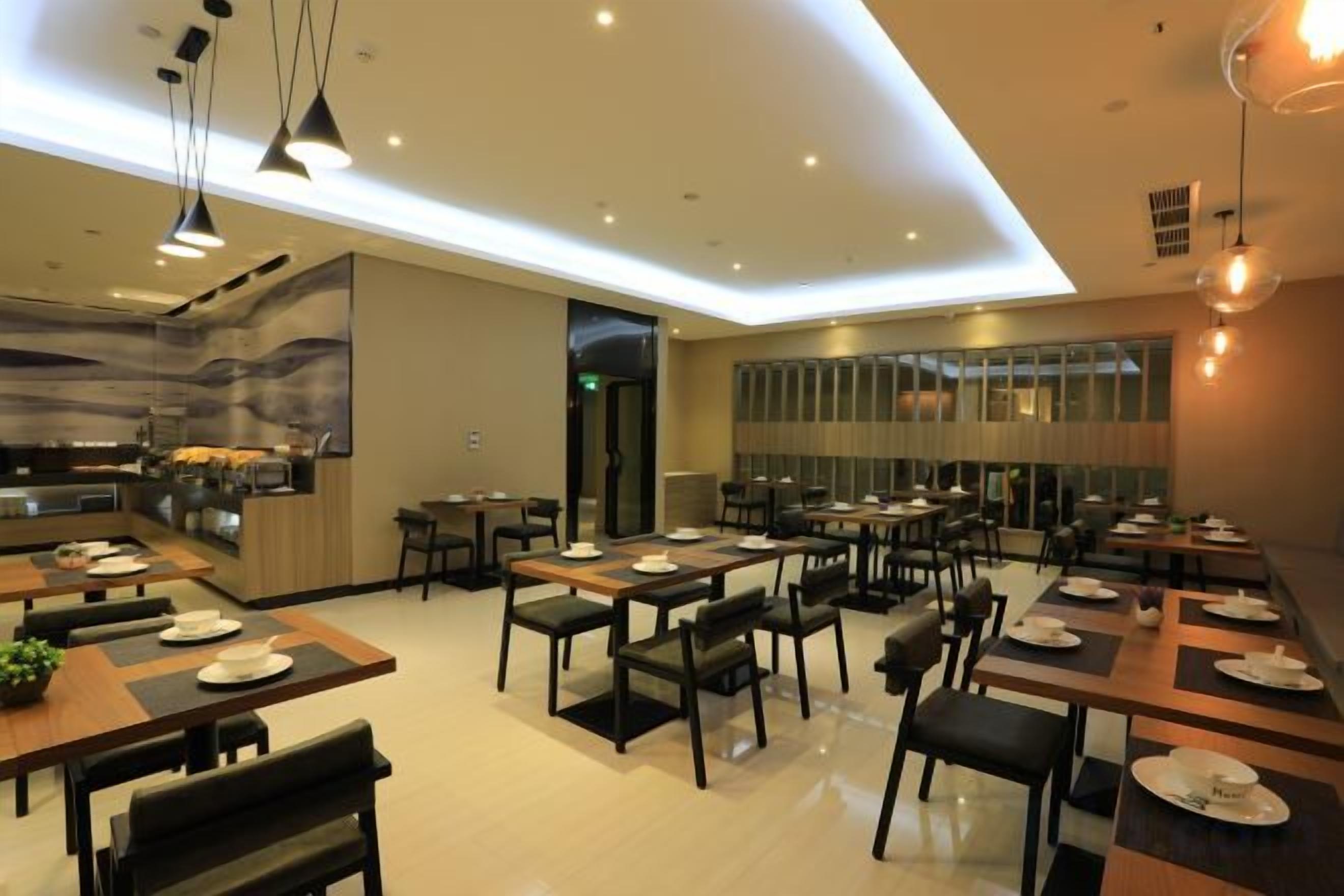 A modern dining area with multiple tables and chairs, a kitchen in the background, and pendant lighting.
