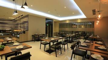 A modern dining area with multiple tables and chairs, a kitchen in the background, and pendant lighting.