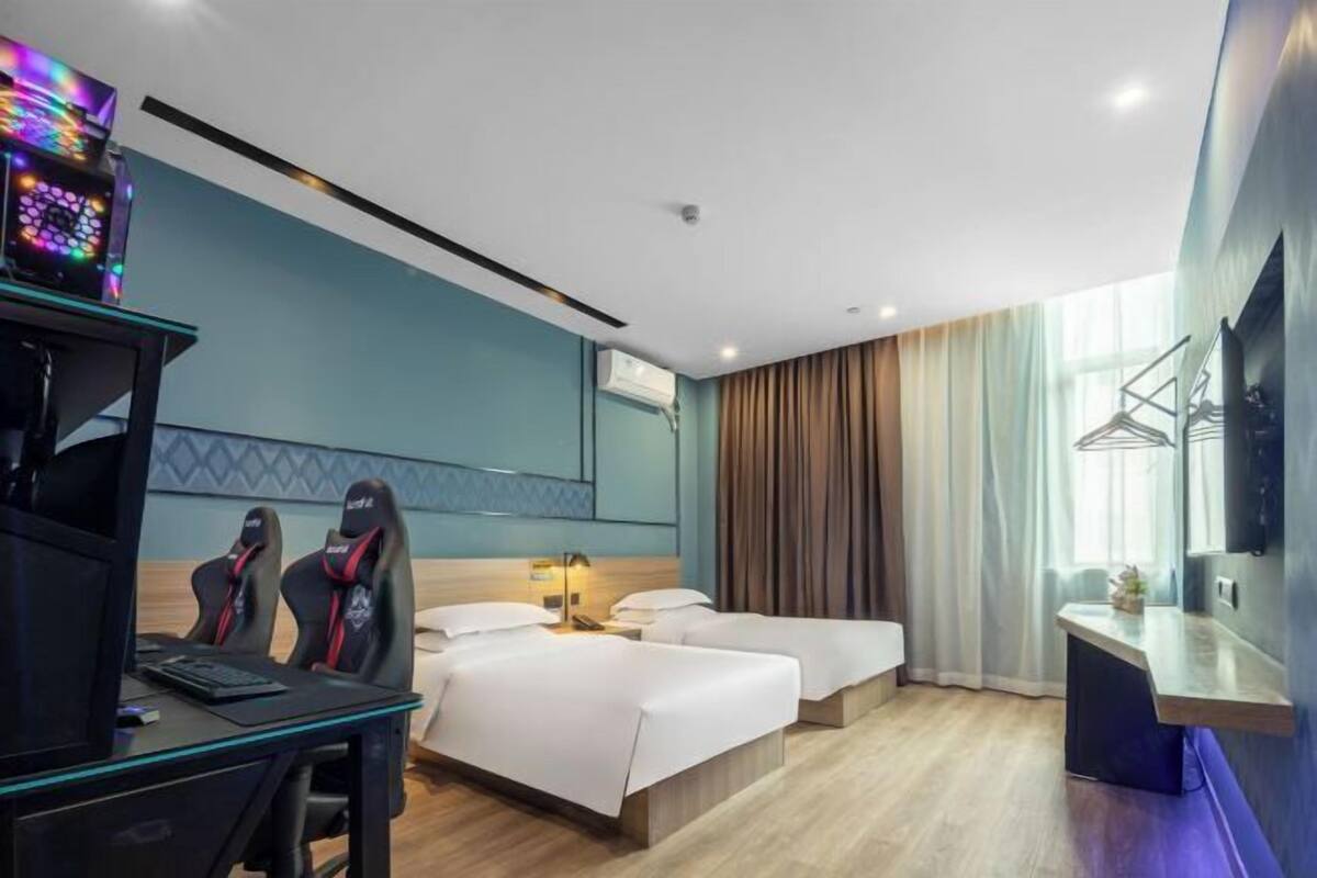 A hotel room with a desk, two gaming chairs, a bed, a bedside table, a TV, and a window with curtains.