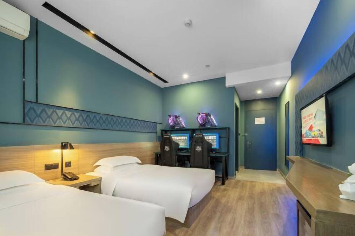 A hotel room with two beds, a desk with two monitors, and a wall-mounted TV.