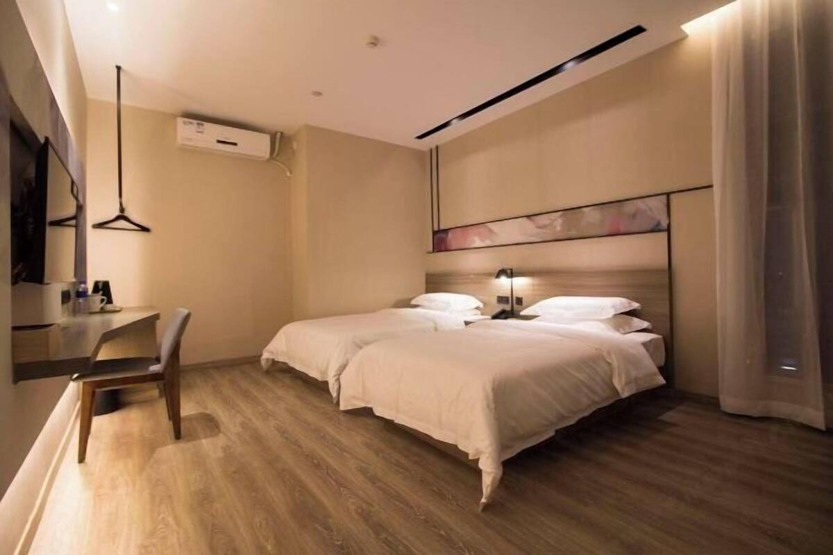 A modern hotel room with a wooden floor, two beds, a desk with a chair, an air conditioning unit, and a painting on the wall.