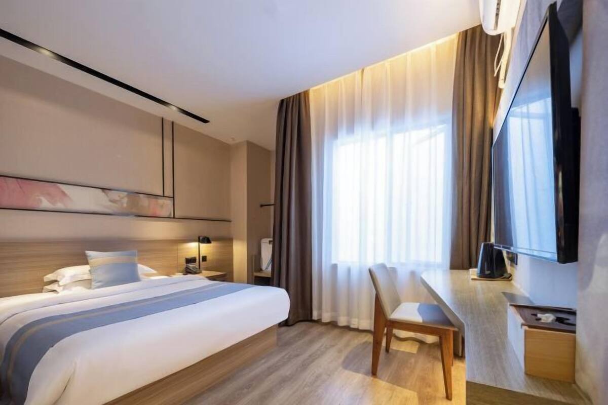 A hotel room with a bed, a desk, a chair, a TV, and a large window with curtains.
