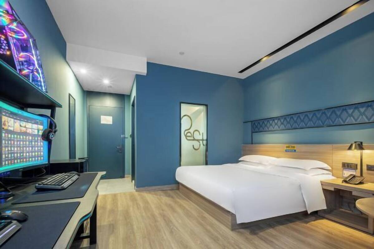 A modern hotel room with a bed, desk, computer, and a neon sign on the wall.