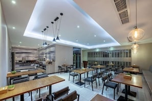 Un restaurant moderne doté de nombreuses tables et chaises, d’un espace cuisine et d’un plafond haut avec des luminaires encastrés.