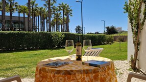 An outdoor table with two wine glasses, a bottle of wine, and two books, set against a backdrop of a well-maintained lawn and tall palm trees.