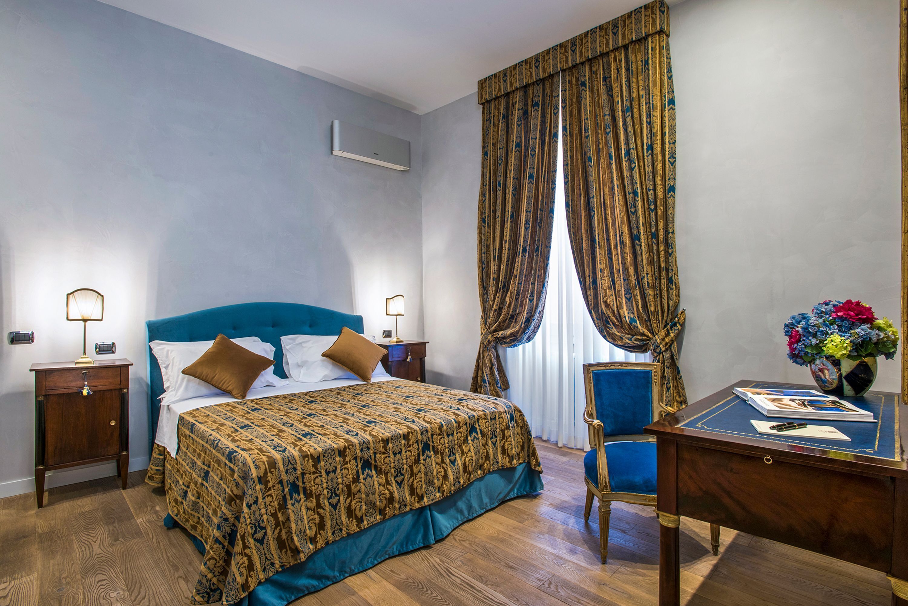 A bedroom with a large bed, a desk with a chair, a nightstand, and a vase of flowers.