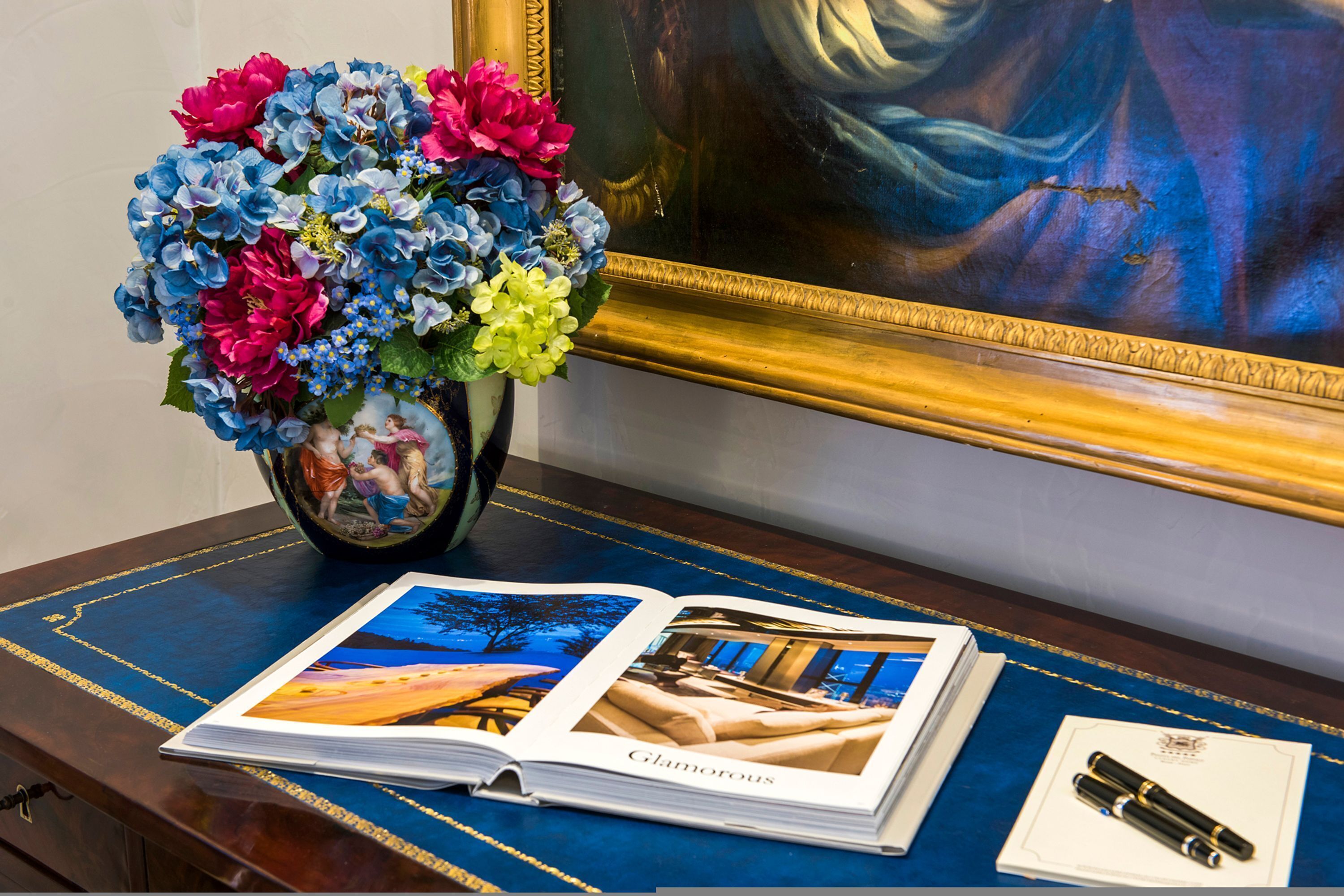 Un bureau sur lequel est posé un livre ouvert à une page intitulée « Glamour », un vase rempli de fleurs et un tableau encadré.