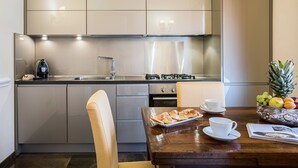 A modern kitchen with a dining area, featuring a stainless steel refrigerator, gas stove, and a wooden dining table with croissants and a fruit bowl. - Alta Luxury Apartments - Oca Apartment (Rome)