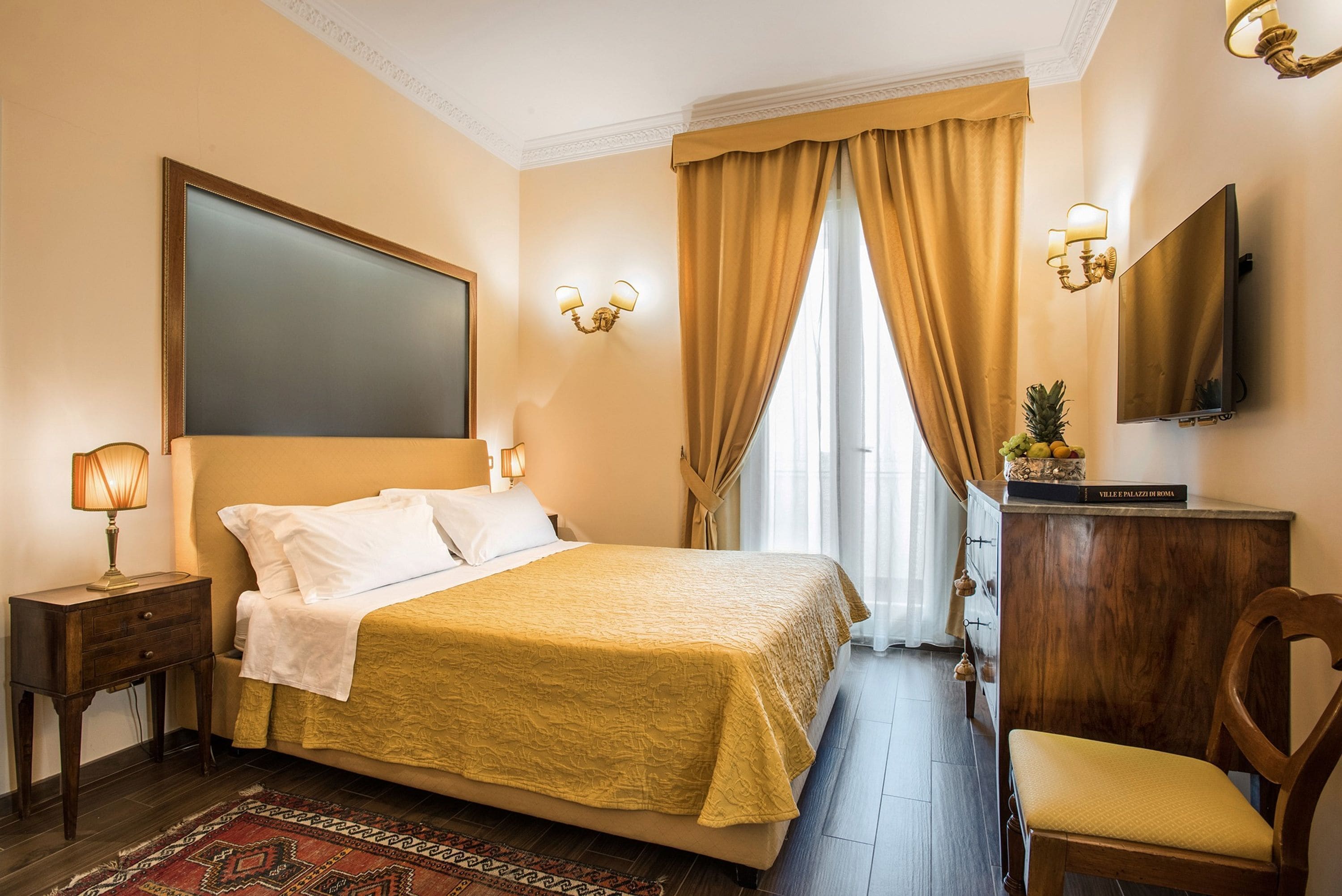 A hotel room with a large bed, a wooden nightstand, a desk with a lamp, a chair, a television, and a window with curtains.