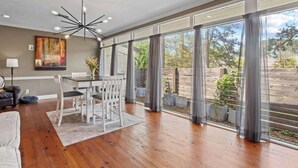 A spacious living area with a dining table, white chairs, a modern chandelier, and large windows offering a view of the outdoors.