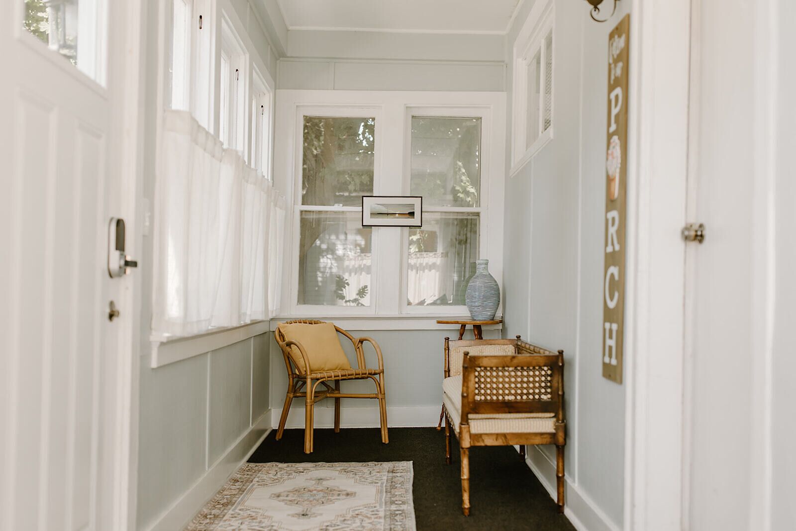 A narrow hallway with white walls, a window with a picture frame, two wicker chairs, a small table with a vase, and a rug on the floor.